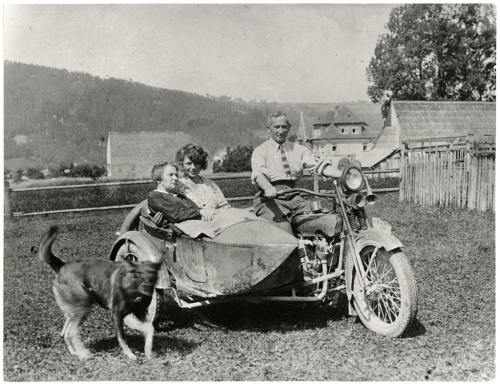 Józef Oppenheim na motorce Harley-Davidson. V sajdkáře sedí Rudá Wanda a Mikucka, okolo běhá pes Rolf.